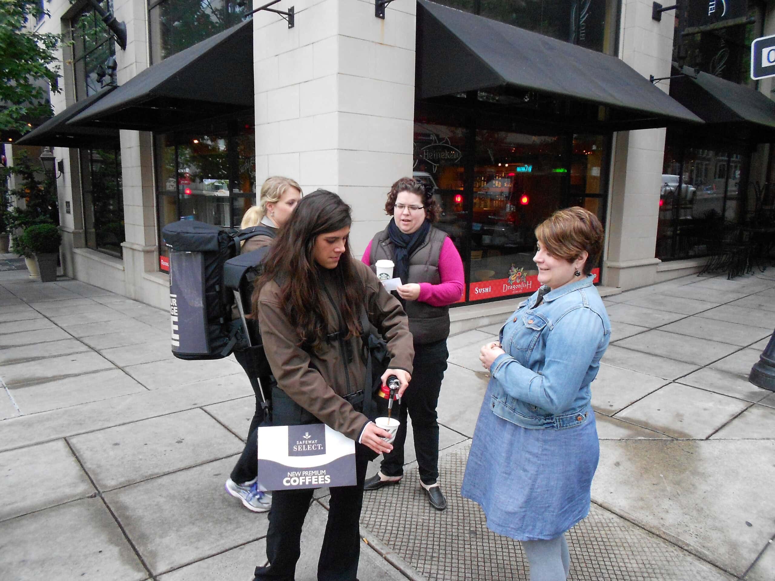 Street marketing activation where a person wearing a backpack coffee dispenser hands out coffee samples to pedestrians on a city sidewalk outside storefronts.